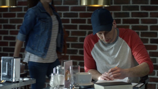 Jeune homme avec casquette assis à une table d'un café dans publicité Salut les amoureux de la Chambre des Notaires réalisée par Yan Giroux chez Alt productions